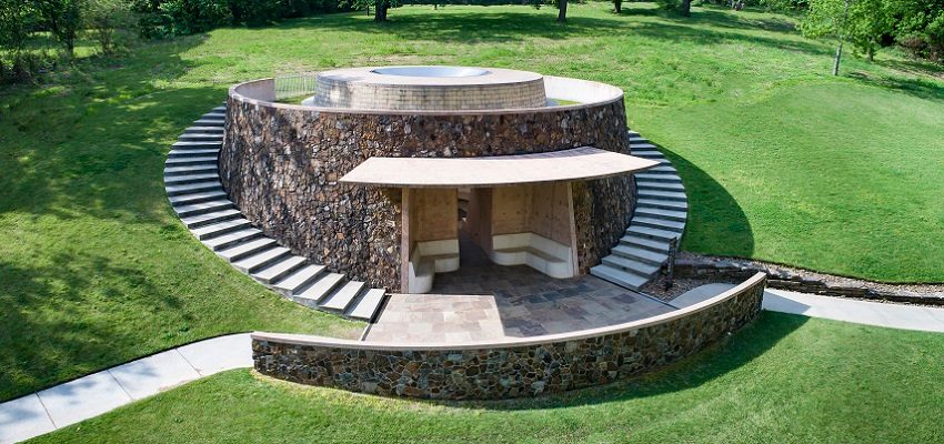 Circular stone structure with steps on grassy landscape, central open space, modern design.