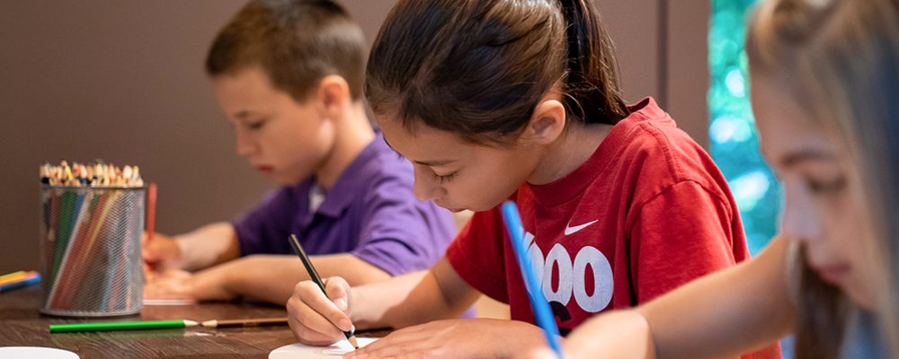 Children drawing and writing at a table with colored pencils and art materials.