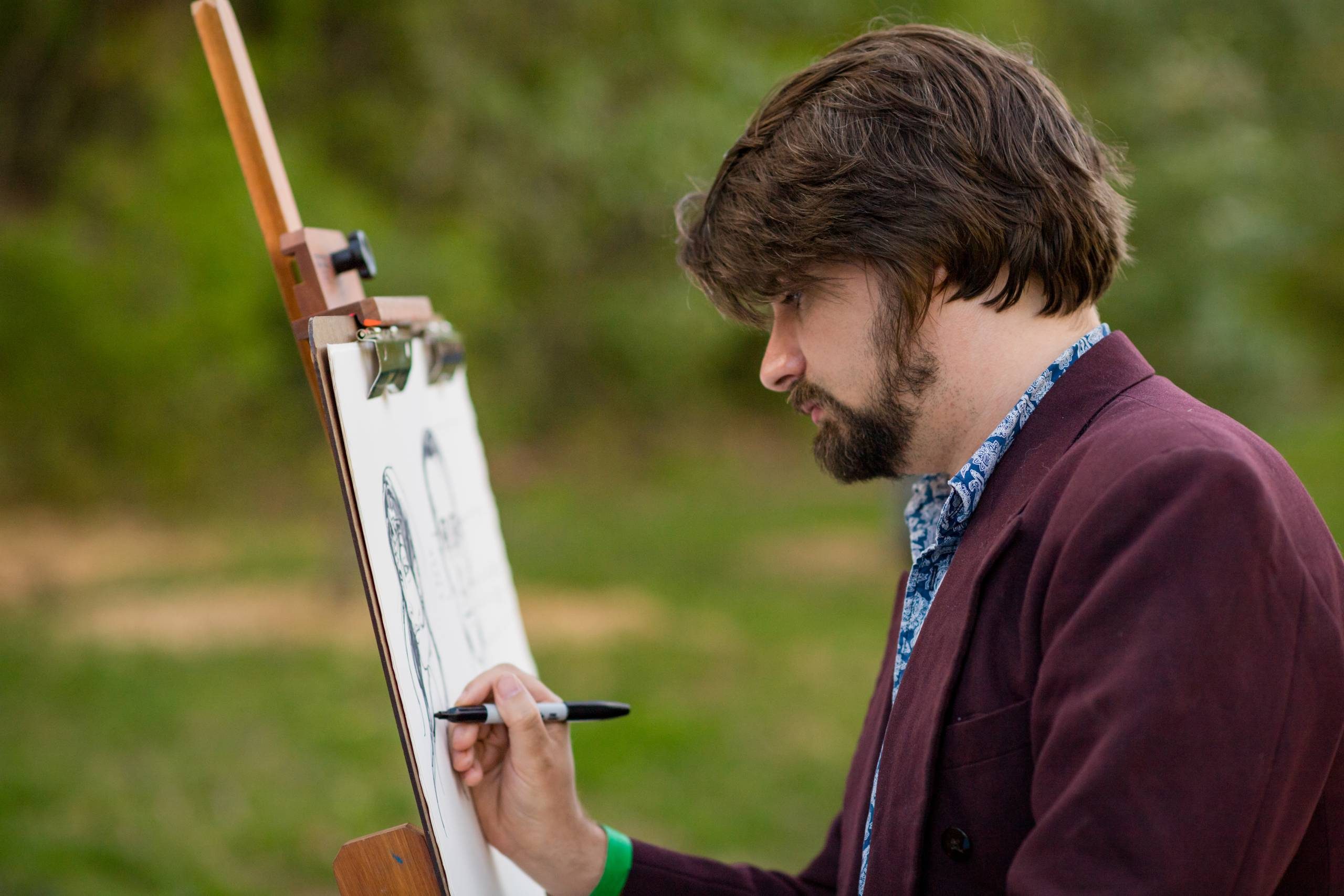 A bearded man in a cardigan draws on a canvas outdoors
