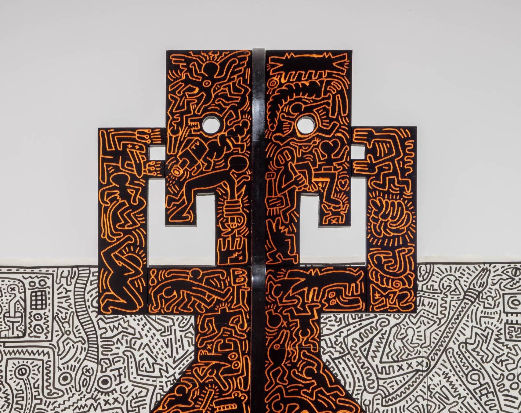 Black sculpture with orange graffiti-style figures stands before a black and white mural.