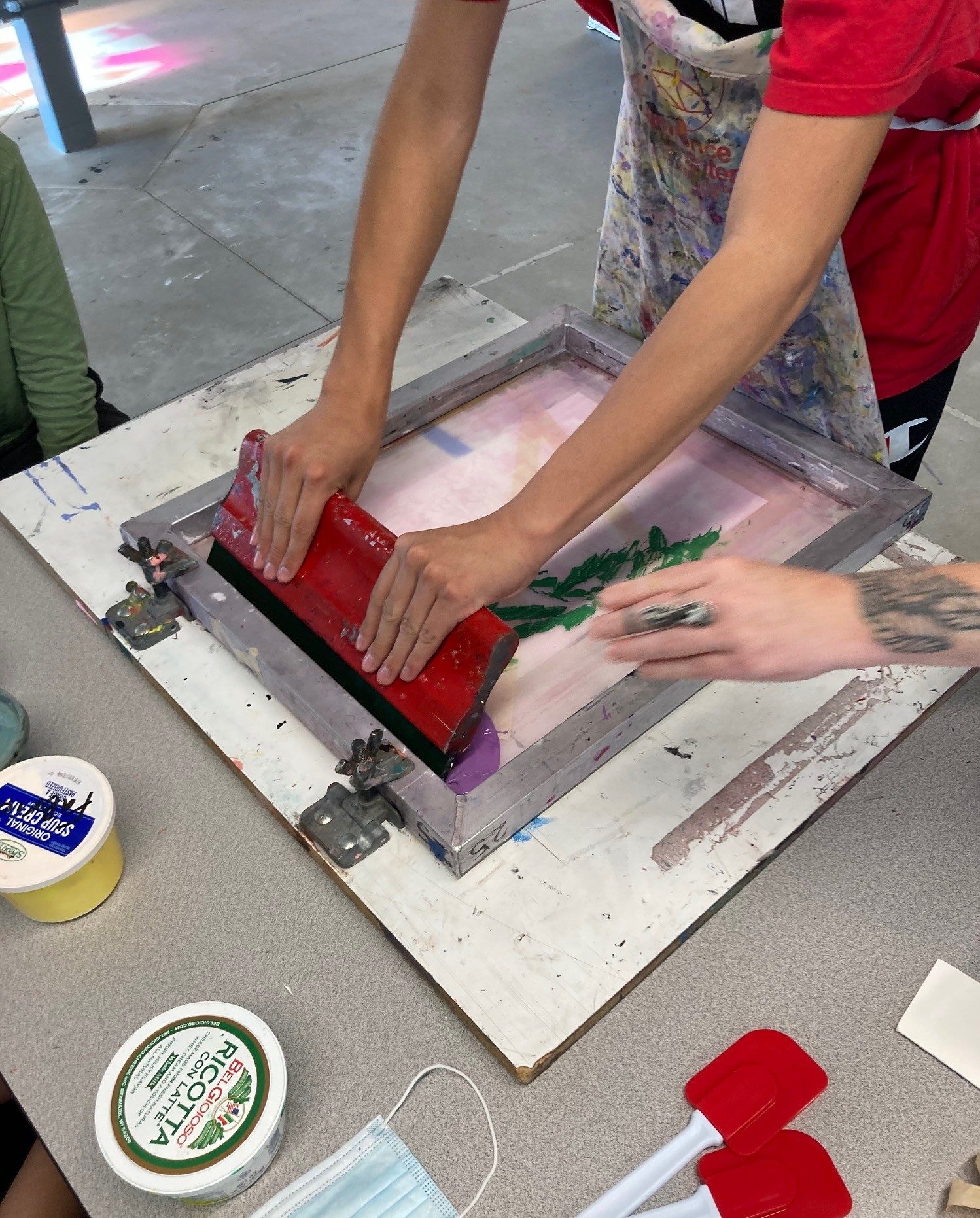 Hands using a red squeegee to spread green and purple ink through a screen printing frame.