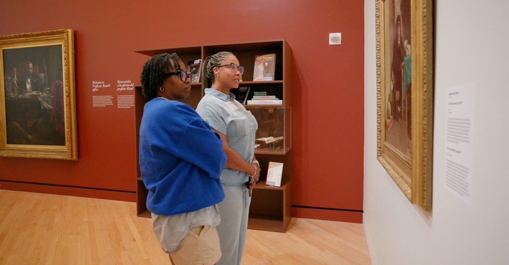 Two people standing in an art gallery with wooden floors and reddish-brown walls viewing paintings.