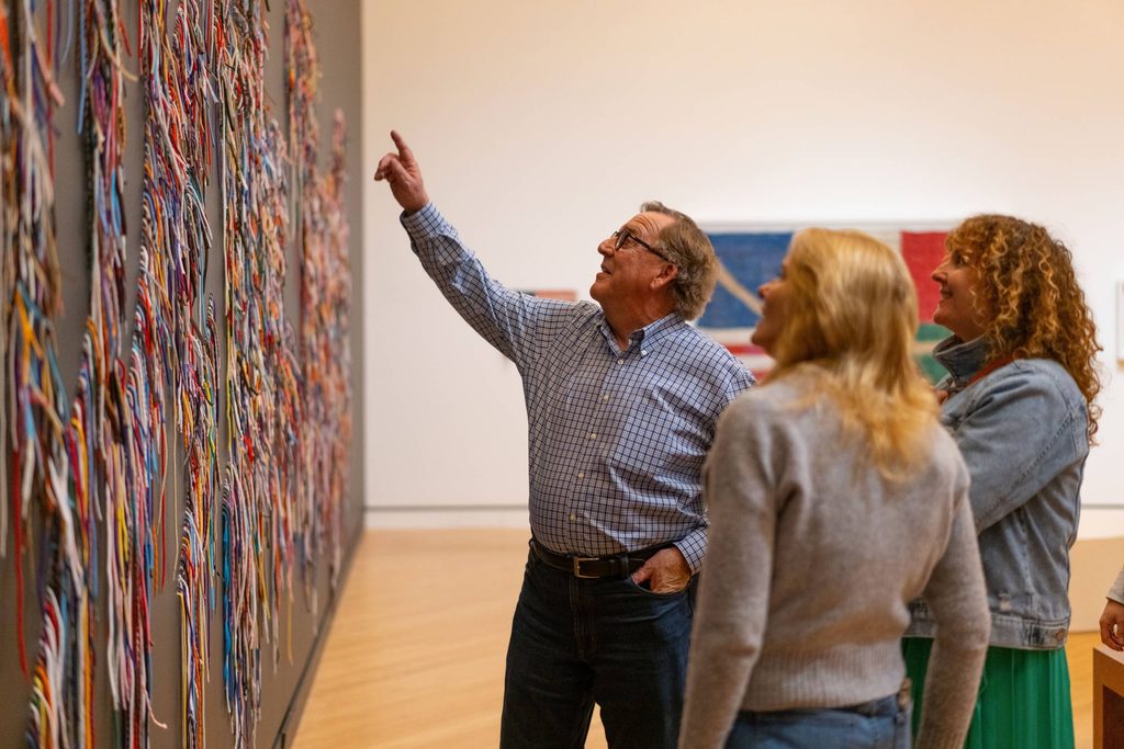 Three people view and discuss a colorful string art installation in a modern gallery.